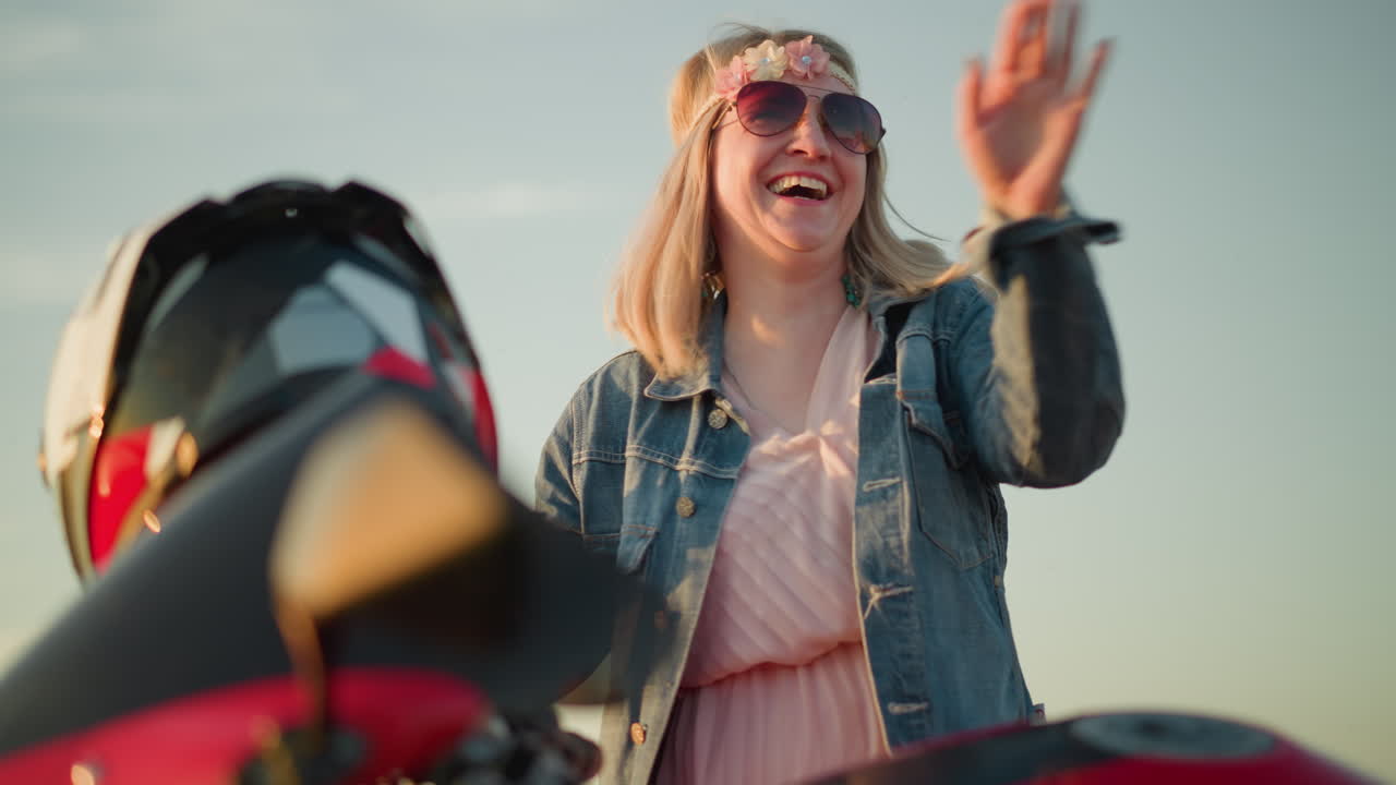 A cheerful woman wearing sunglasses and a flower headband is dancing with her hands raised and smiling while standing next to a red motorcycle in a grassy field