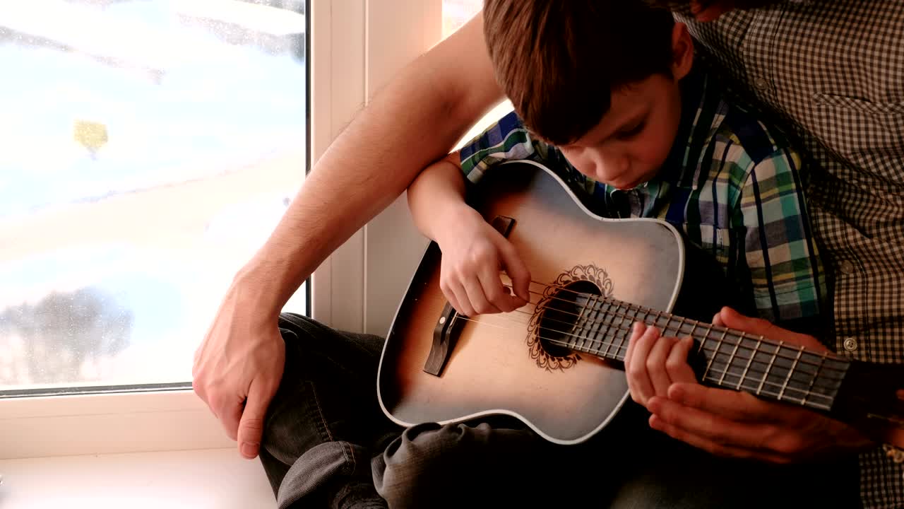 papá le enseña a su hijo a tocar la guitarra, sentado en el alféizar de la ventana.
