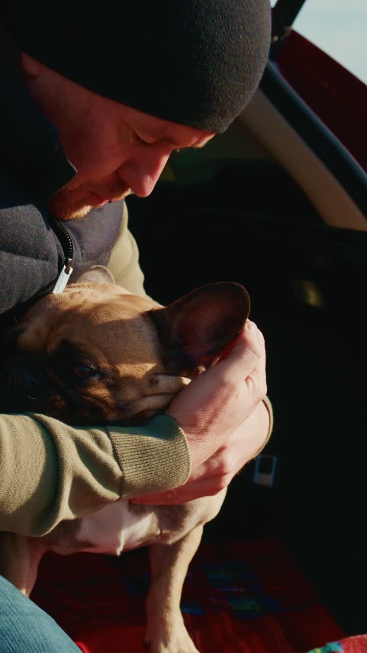 Un legame commovente: una persona conforta il suo bulldog francese sul sedile posteriore di un'auto, mettendo in mostra l'amore e il legame tra l'uomo e l'animale domestico, sullo sfondo di un pittoresco paesaggio esterno