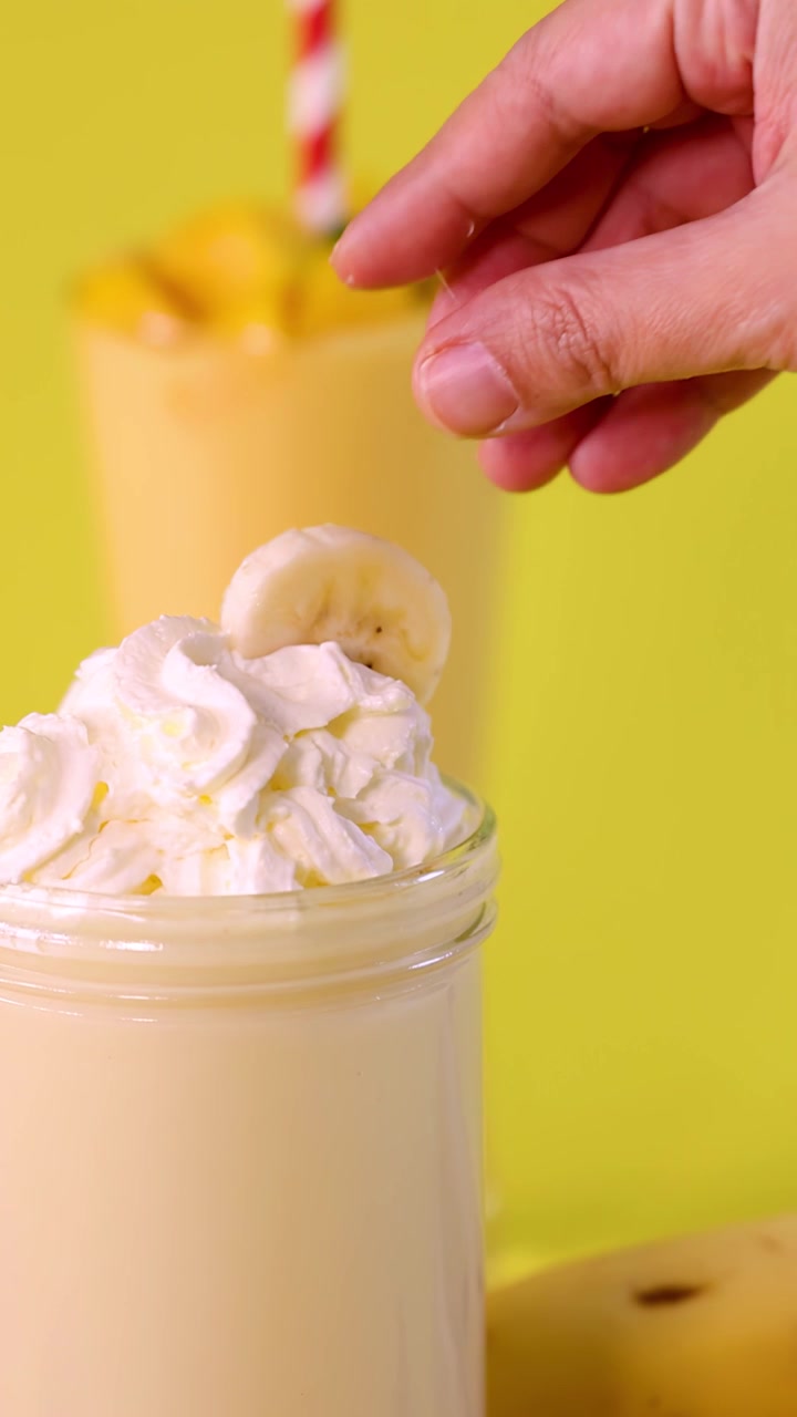 Una mano posiziona una fetta di banana su un frullato alla banana con panna montata, sfondo giallo brillante, illuminazione da studio