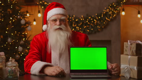 Der-Weihnachtsmann-Hält-Einen-Laptop-Mit-Einem-Grünen-Bildschirm,-Während-Er-In-Der-Weihnachtsdekoration-Sitzt.-Hochwertiges-4K-Filmmaterial