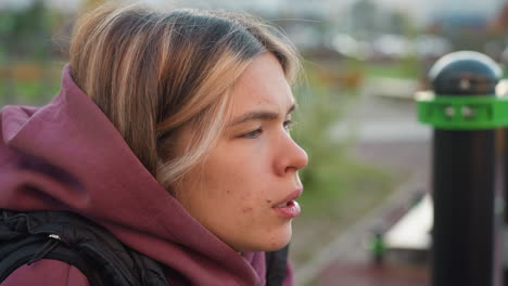 mid shot of fitness trainer leaning on bar after intense outdoor workout, wearing maroon sweatshirt, catching breath with visible effort, muscular arms resting on metal bar, blurred building backdrop