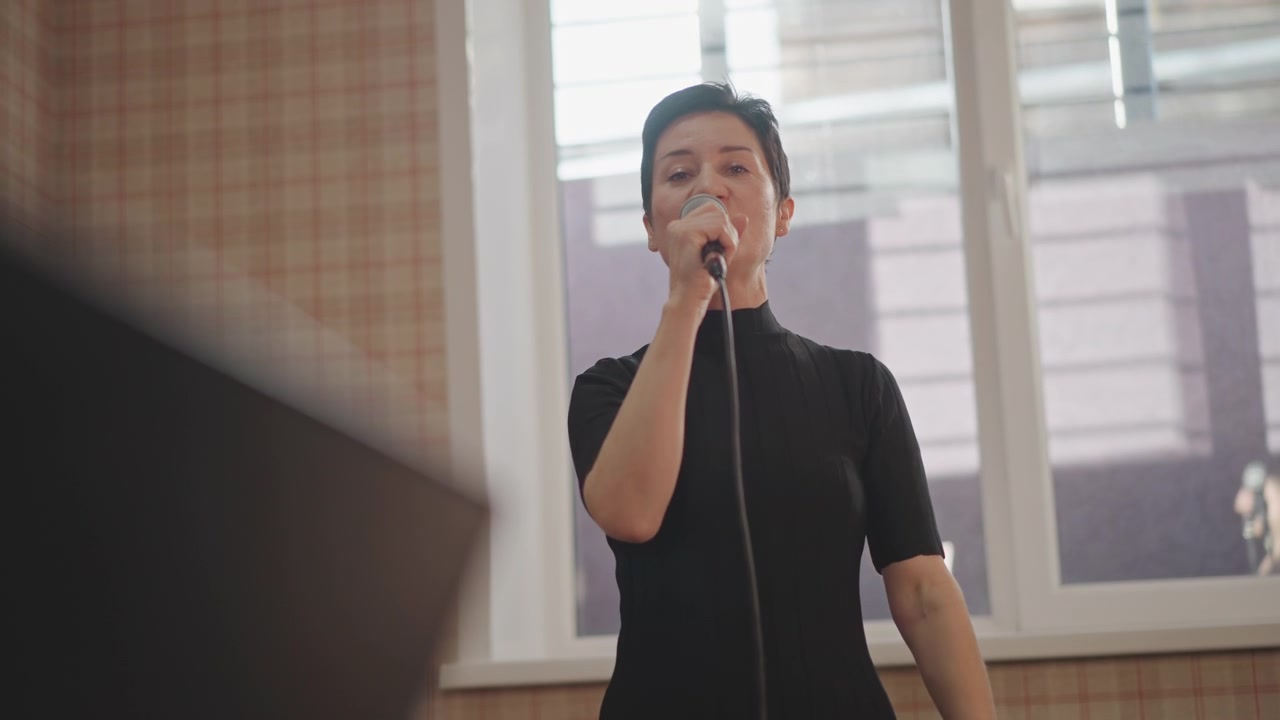 Performer synger med udtryksfulde håndbevægelser, nodestativ i forgrunden, slørede noder, engagerende smil, fortællende frasering, afslappet øvestemning, naturligt lys, draperet mikrofonledning