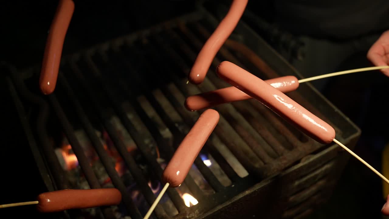 Des hot-dogs rôtissant sur des brochettes sur un barbecue la nuit