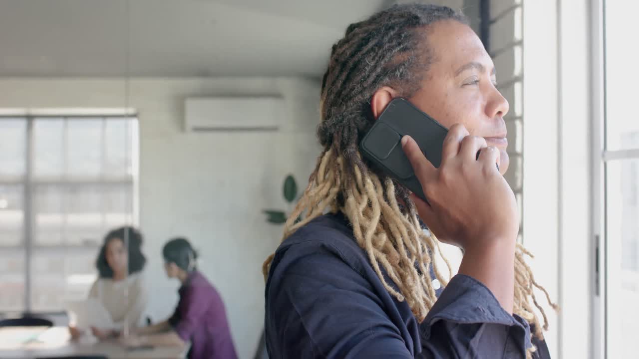 Uomo creativo birazziale felice che parla al telefono e colleghi che parlano sullo sfondo, slow motion
