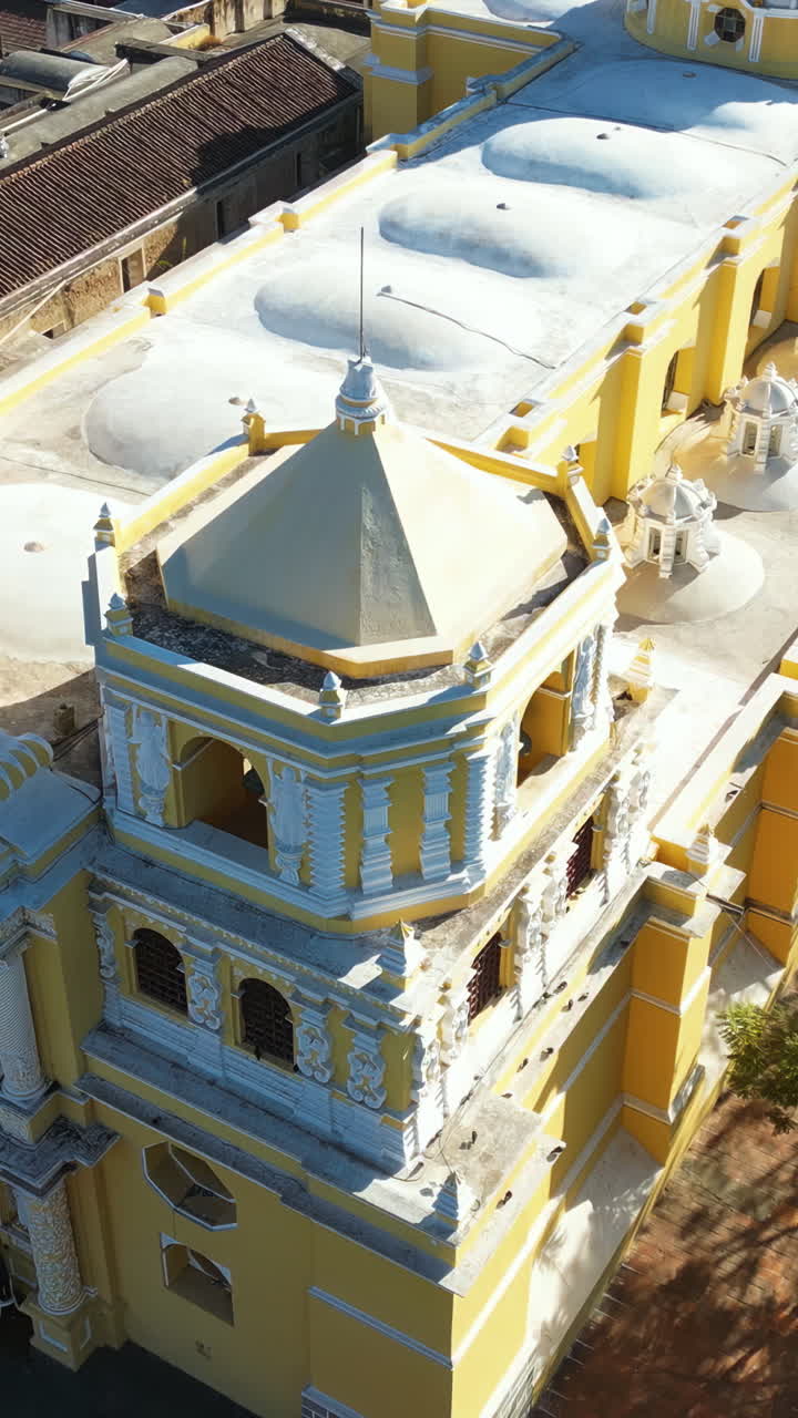 Luftfoto fra drone af den ikoniske gule La Merced Kirke i Antigua Guatemala, omgivet af koloniale bygninger og palmetræer. Vertikal.