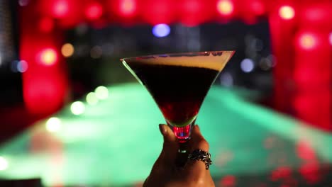 a hand swirls an espresso martini in a cocktail glass at a vibrant bar with neon red and green lighting, shallow depth of field, and smooth camera movement