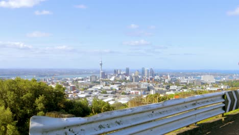 A-dolly-in-shot-of-the-Auckland-skyline-in-New-Zealand,-viewed-from-a-mountain-path-with-two-birds-passing-by-on-a-clear-afternoon-with-a-blue-sky