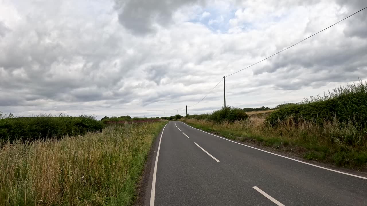 Car driving along winding rural road bordered by hedges and fields under cloudy daylight sky