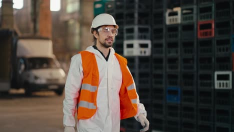 Un-Hombre-Con-Barba-Y-De-Piel-Morena,-Con-Un-Uniforme-De-Protección-Blanco,-Un-Casco-Blanco-Y-Un-Chaleco-Naranja,-Camina-Junto-A-Cajas-Negras-Y-Amarillas-En-Una-Enorme-Planta-De-Procesamiento-Y-Clasificación-De-Residuos.