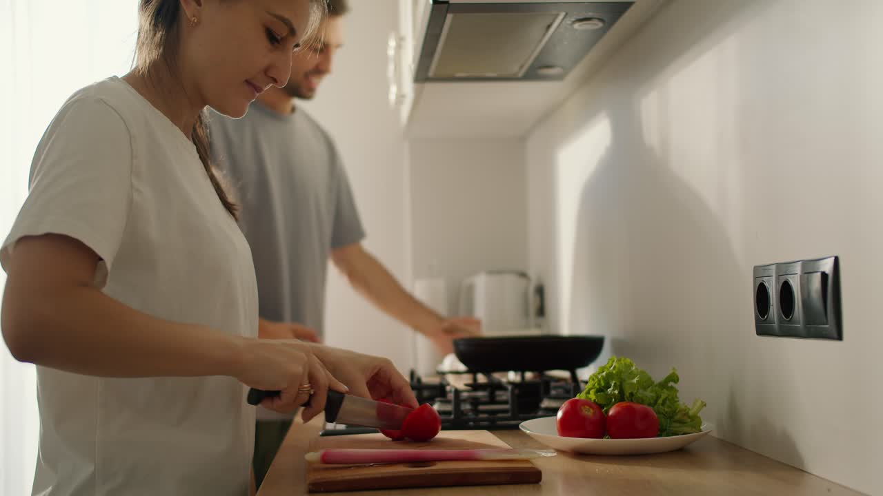 una chica morena con una camiseta blanca está preparando una ensalada matutina y su marido, un tipo con una camisa gris, habla con ella y cocina algo en la estufa de la cocina por la mañana