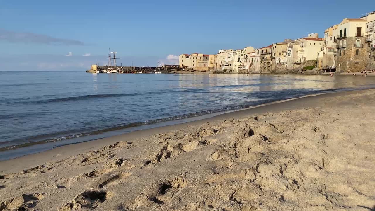 Ampia inquadratura statica della costa di Cefalù, Sicilia, che cattura le calme acque mediterranee che raggiungono dolcemente la riva sabbiosa in primo piano. Sullo sfondo, il centro storico si erge accanto al mare