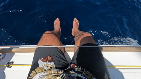 a man wearing shorts and a tropical shirt sits on boat hanging his legs over the side as the water passes underneath