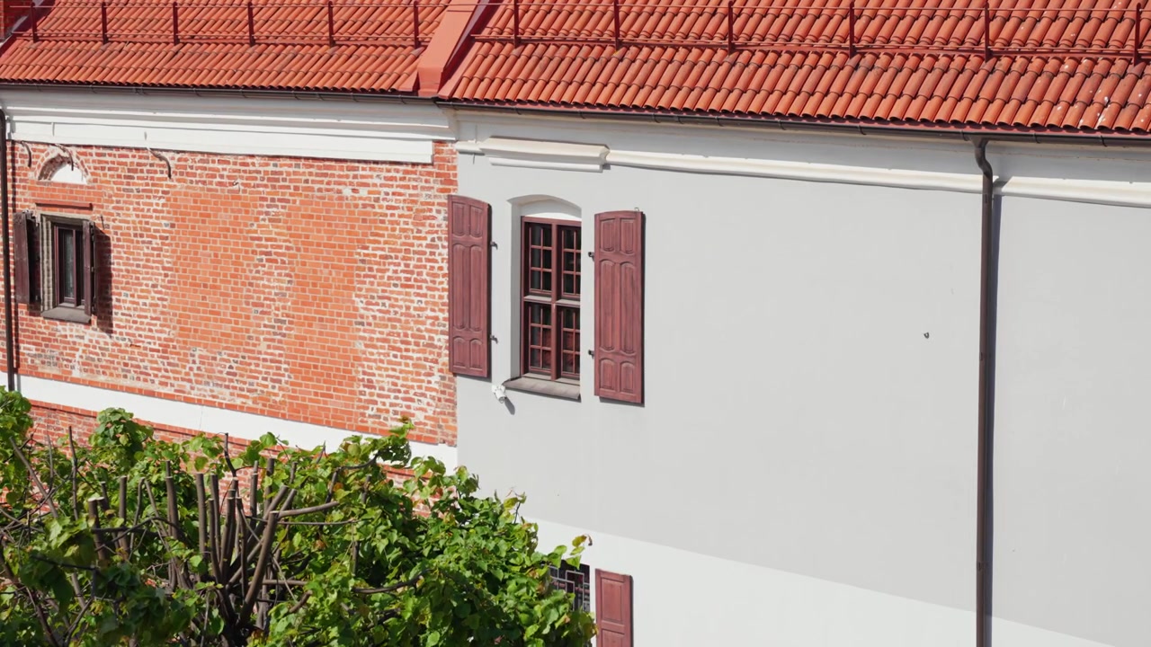 Jasné sluneční světlo zalévá klasickou cihlovou a štukovou budovu, představující červenou taškovou střechu, texturované zdivo, dřevěné okenice a bujnou zeleň v klidném, stabilním širokoúhlém záběru