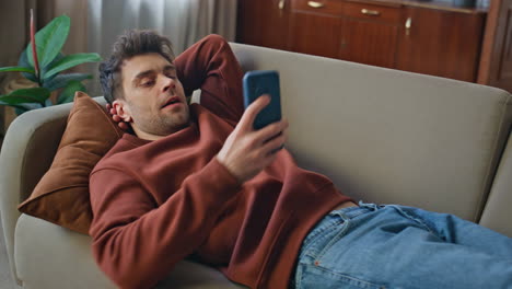 Resting man lying couch holding smartphone closeup. Young guy looking cellphone