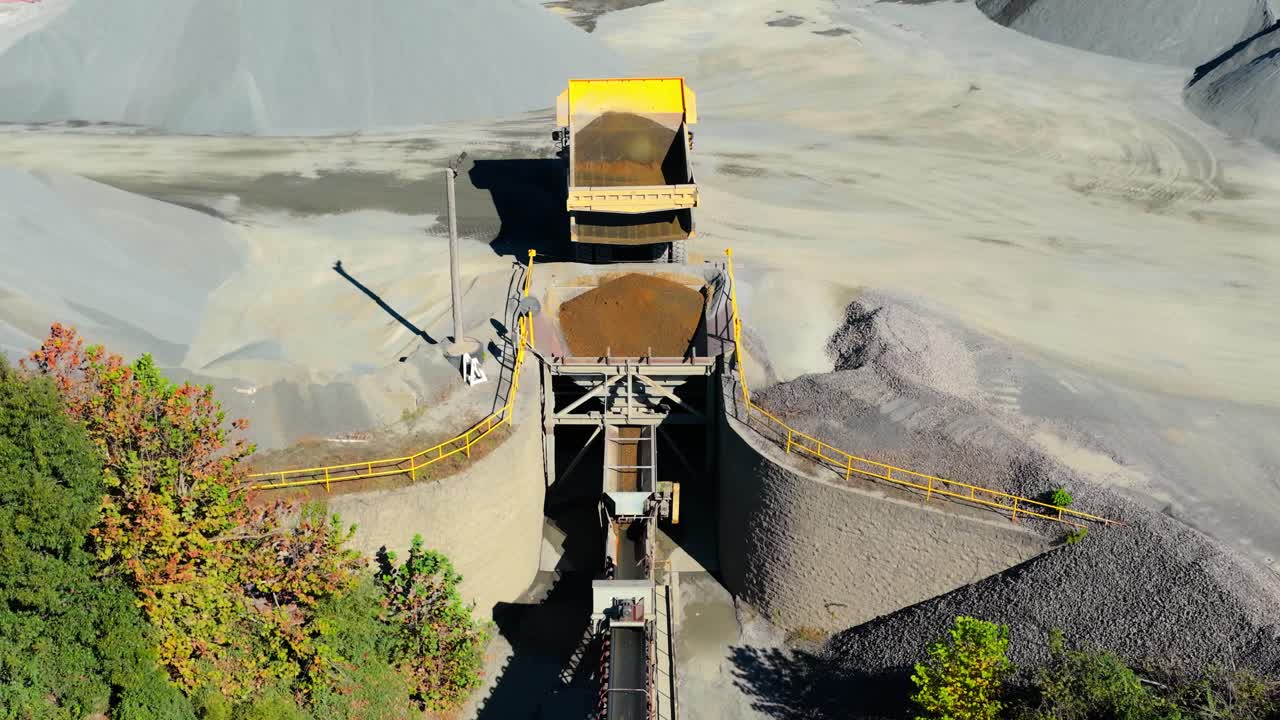 Dump truck positioned above hopper emptying and filling it with red aggregate in quarry, aerial approach