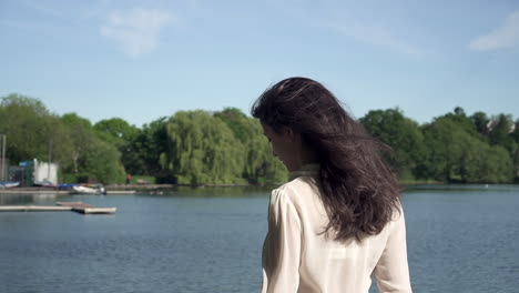 Portrait-of-a-fashionable-and-beautiful-Italian-model-walking-in-a-tranquil-park-near-a-lake,-wearing-a-transparent-shirt,-looking-around-and-having-fun-in-London,-United-Kingdom