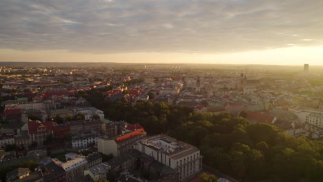 Vuelo-Aéreo-Hacia-Atrás-Sobre-La-Iluminación-De-La-Ciudad-De-Cracovia-Al-Atardecer-Dorado-En-Polonia,-Europa