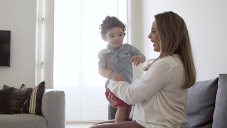 Excited-mother-sitting-on-sofa,-holding-son-in-hands.