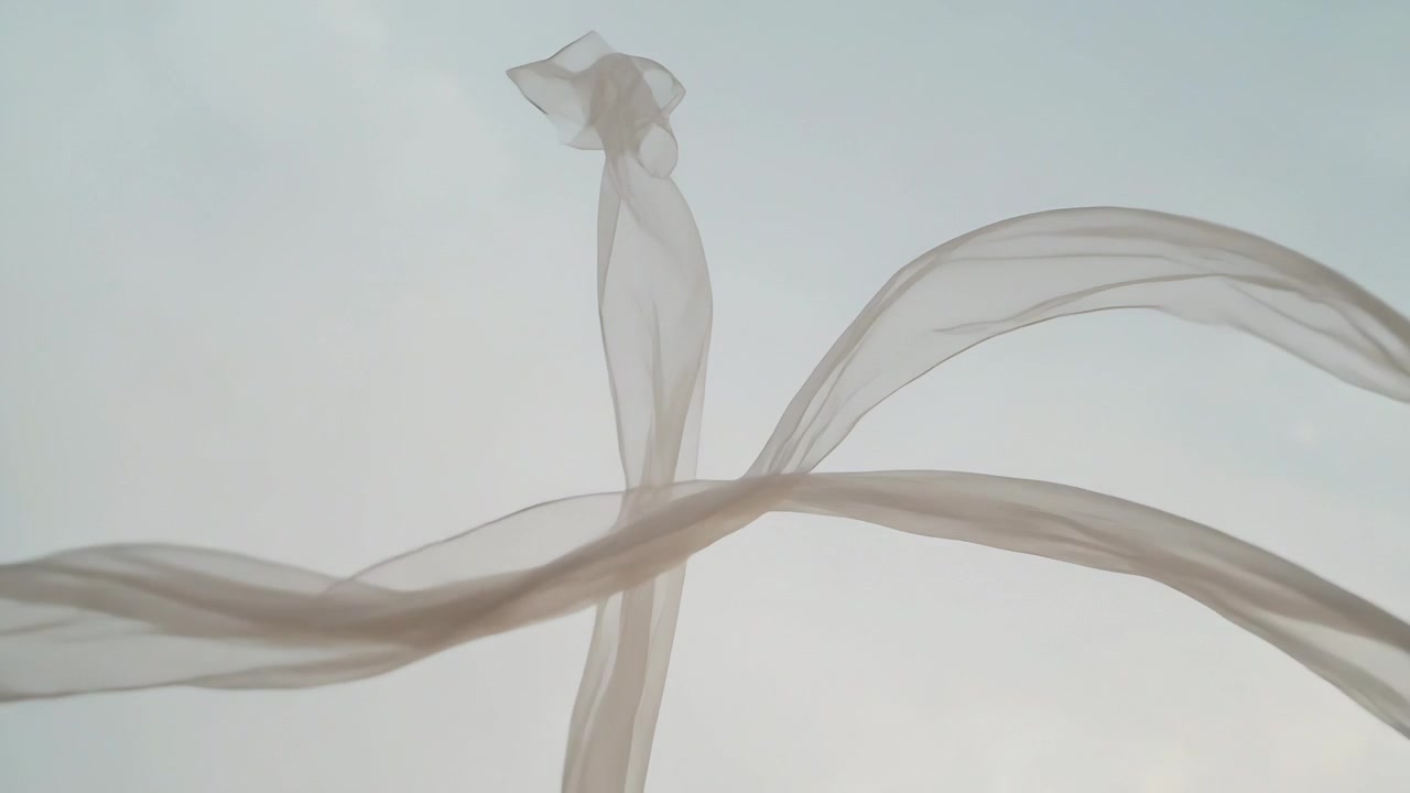 Lange, transparente Stoffbänder schweben, verdrehen, kreuzen und entfalten sich am hellen Himmel, während eine Brise weht.