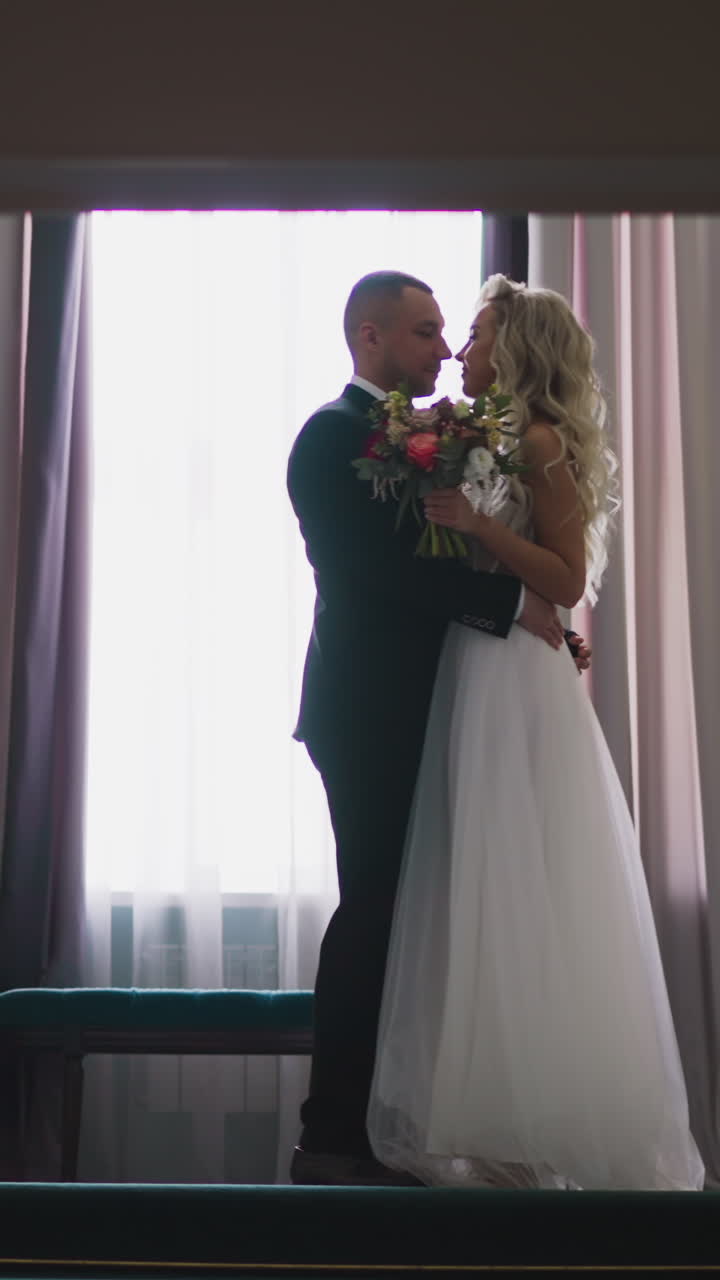 Elegant pretty young bride and happy groom meet halfway on vintage staircase against large window before wedding ceremony slow motion