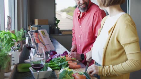 Happy-diverse-couple-cooking-together,-chopping-vegetables-and-drinking-wine-in-kitchen