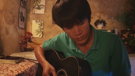 Young-Asian-male-playing-a-guitar-while-on-a-chair-in-a-rustic-room