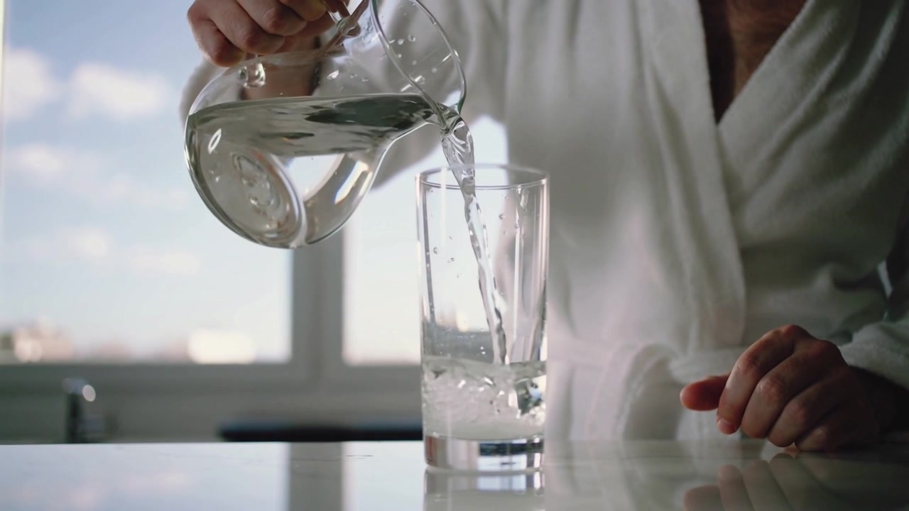 Ein Mann in weißem Gewand kippt einen Krug und gießt Wasser in ein Trinkglas an der Küchenzeile.