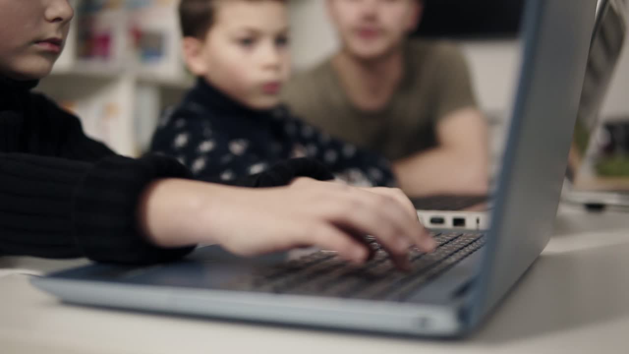 Kid's arms typing somethingon laptop keyboard with two unrecognizable figures sitting on the background. Programmingfor kids class. Learning how to use technologies and gadgets.
