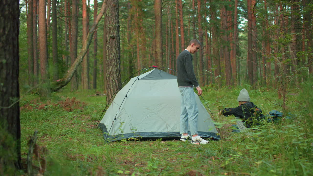 couple playfully exchanging tent peg during camping setup in serene pine forest clearing with soft sunlight filtering through tall trees as they collaborate on outdoor gear assembly