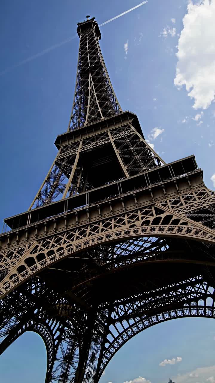 Inquadratura dinamica dal basso verso l'alto della Torre Eiffel su uno sfondo di cielo blu, che cattura il suo intricato lavoro in ferro