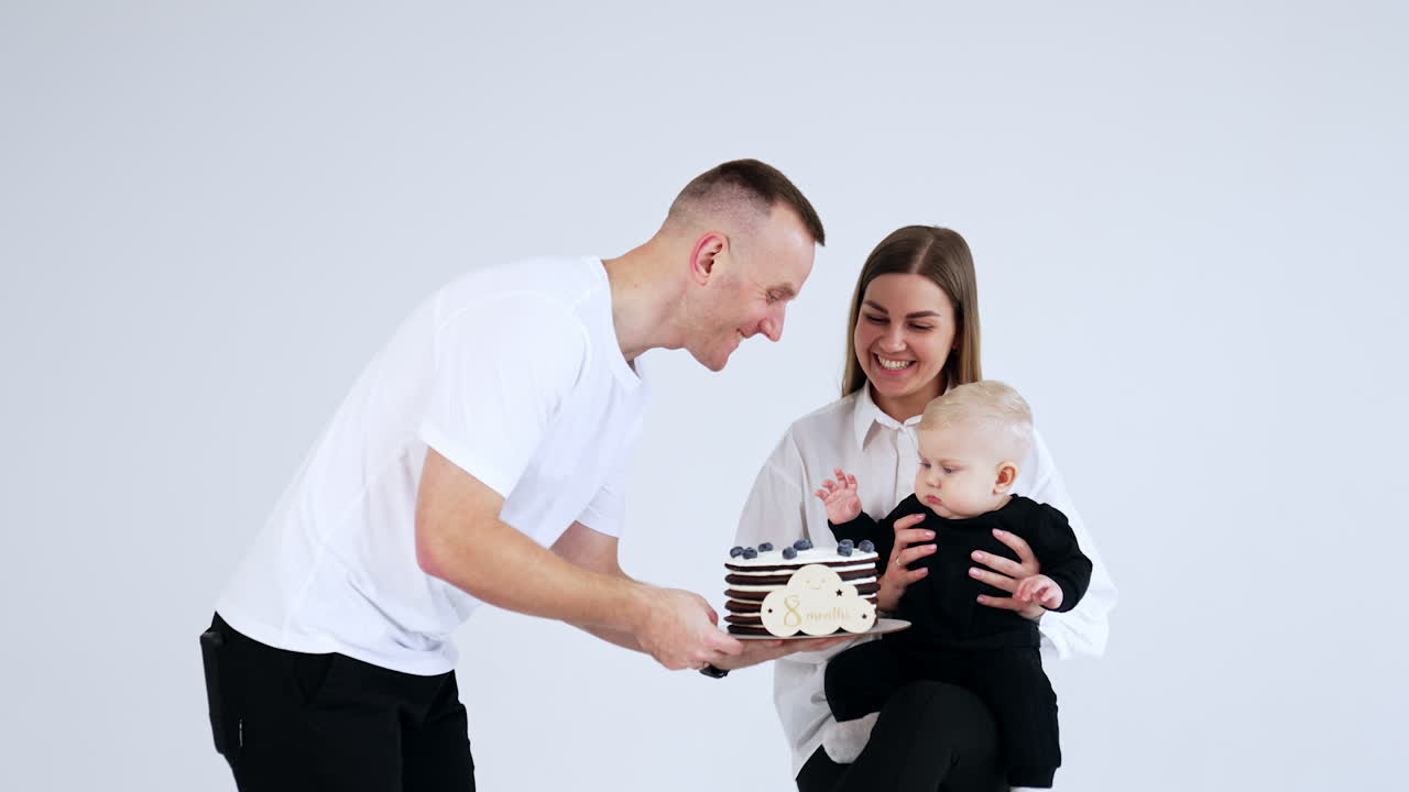 Happy smiling Caucasian mother holding her cute baby boy. Cheerful father comes up bringing a cake and showing the sweet to a child. White backdrop.