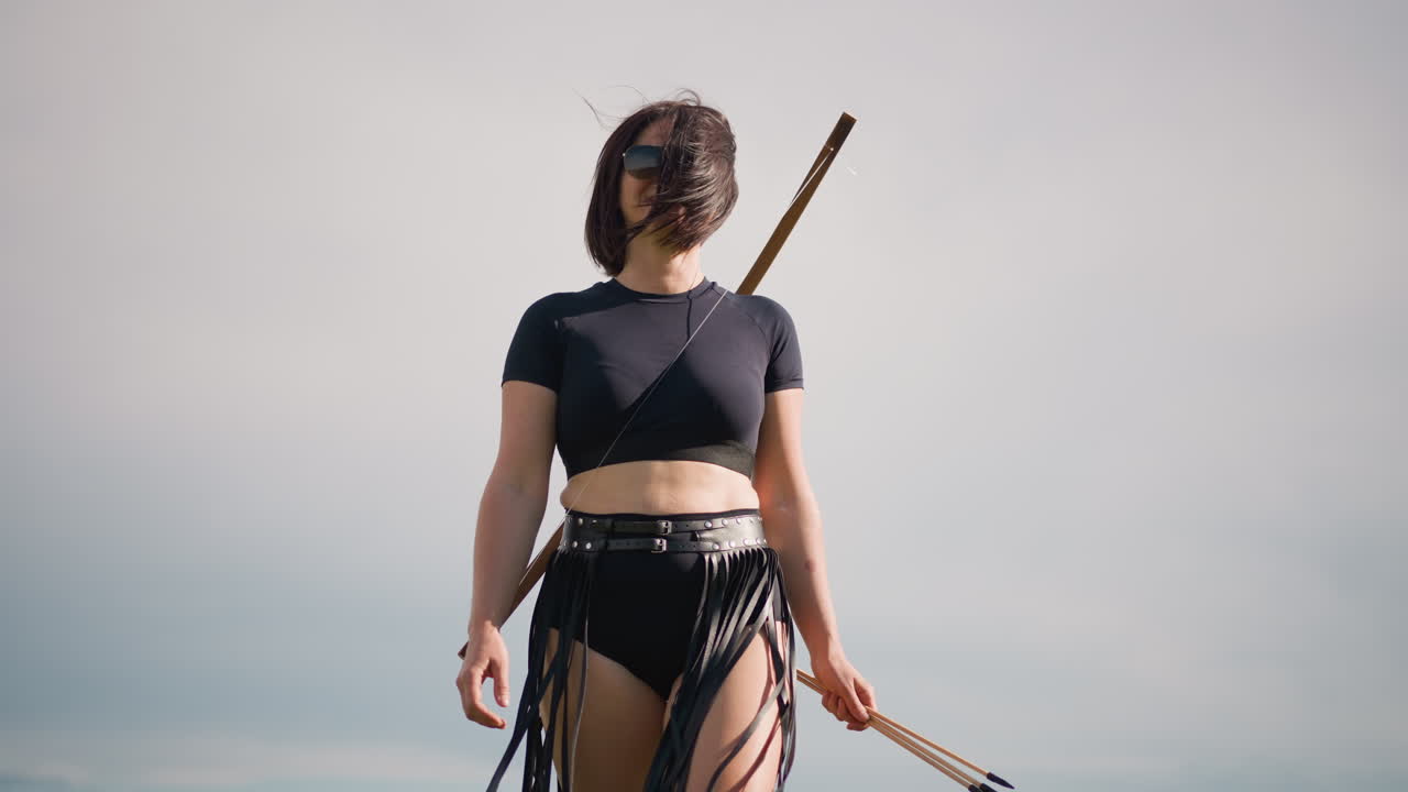 Bílá žena-bojovnice na pozadí zatažené oblohy, kožená sukně s třásněmi a černý crop top, hůl připevněná na zádech, vlasy rozcuchané větrem, nízkoúhlý filmový portrét, osamělá zvědka se zastaví a skenuje obzor, pomalu