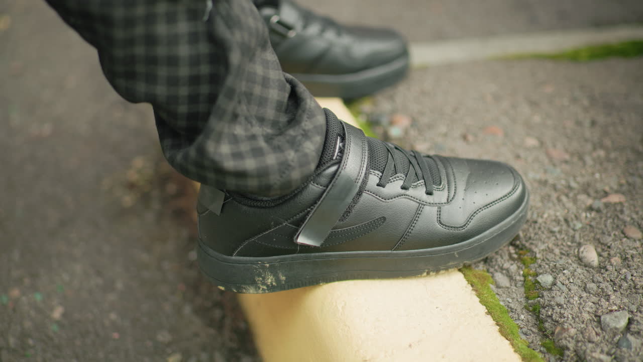 Close up of person legs wearing black sneakers stepping onto yellow pavement curb outdoors, showing casual footwear, street detail, and body movement in an urban environment