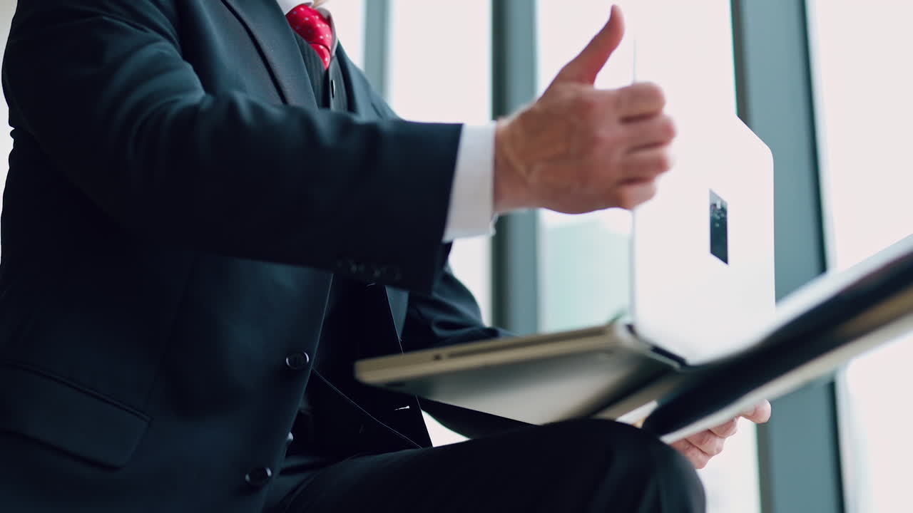 Mature businessman in glasses using laptop. Gray-haired man in costume works with a laptop while sitting at the window in the office.