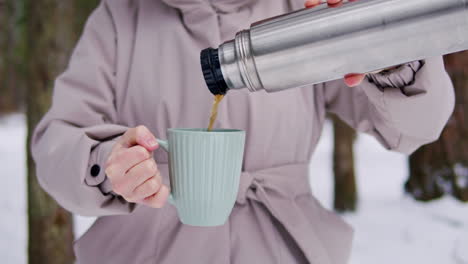 woman pouring hot drink from thermos into mug in winter forest