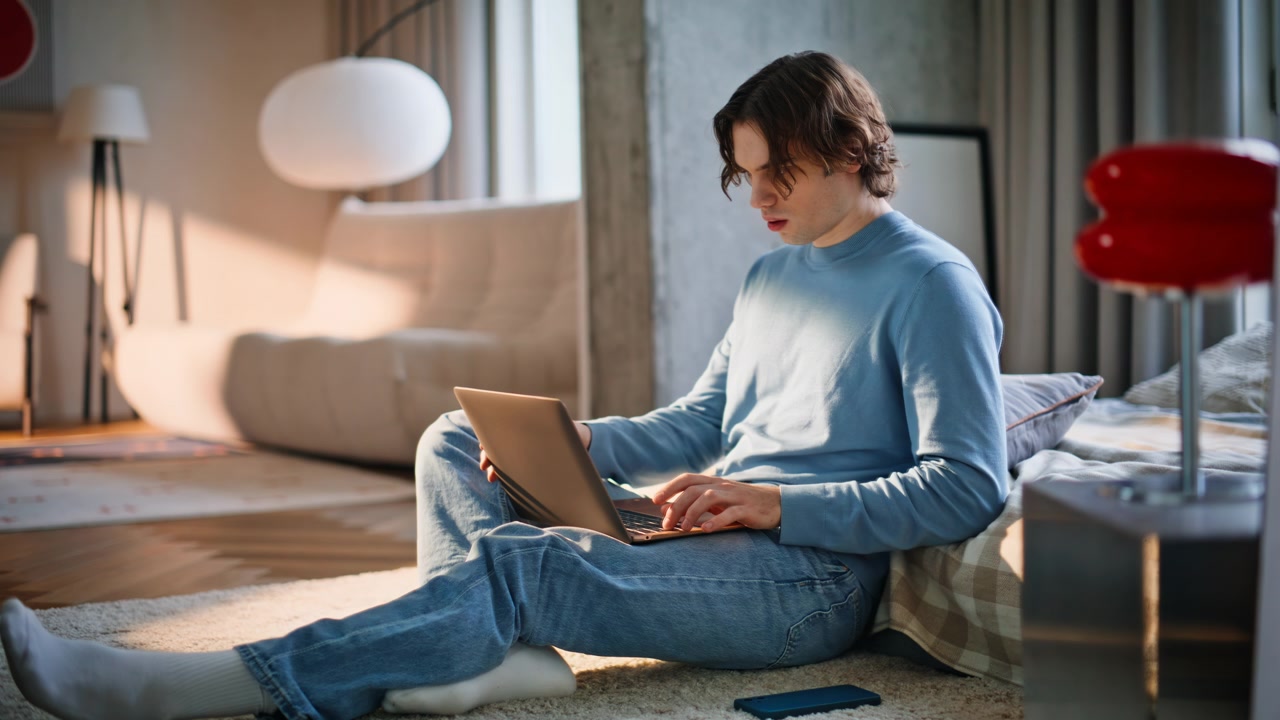 Studying guy sitting floor using laptop in sunny bedroom. Man finishing working
