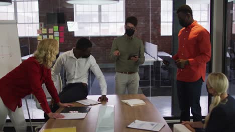 diverse group of business colleagues wearing masks brainstorming in meeting room using tablet
