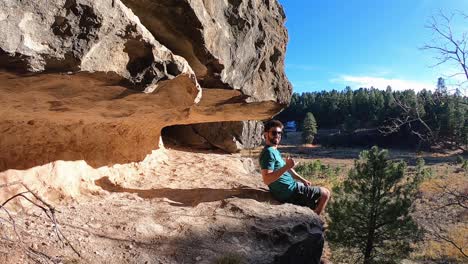 a man sitting on a rock shelf overlooking the coconino forest looks back at the camera and gives a hang loose sign