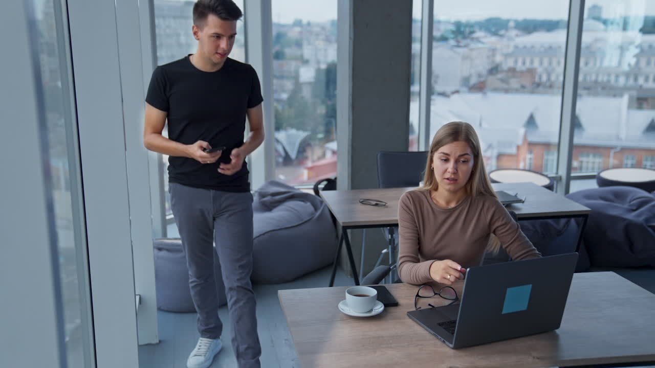 Colleague communication in the office. Female asking for help her male co-worker at computer. Panoramic windows with cityscape at backdrop.