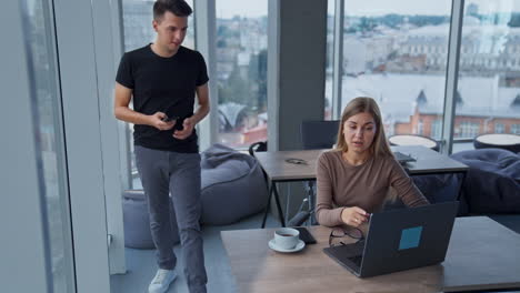 colleague communication in the office. female asking for help her male co-worker at computer. panoramic windows with cityscape at backdrop.
