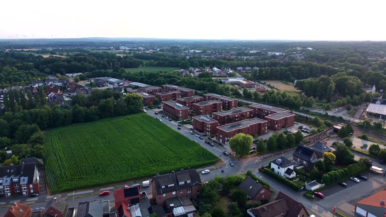 Blocchi di appartamenti in mattoni rossi confinano con un campo agricolo verde accanto a un tranquillo sobborgo tedesco al tramonto dorato; strade alberate e case con tetti rossi si estendono attraverso il quartiere residenziale. Ripresa aerea con drone in avanti