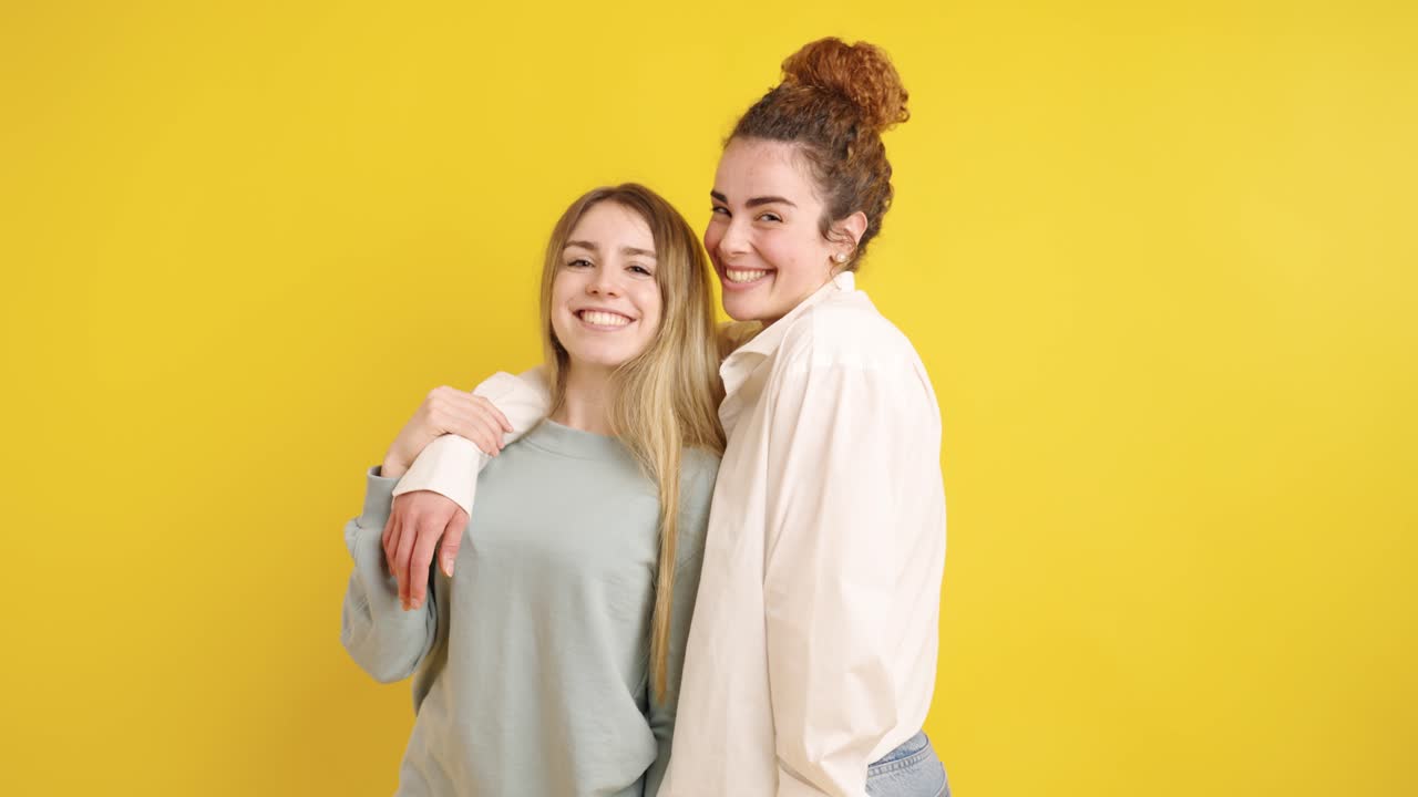 Two young women embracing and smiling on yellow background