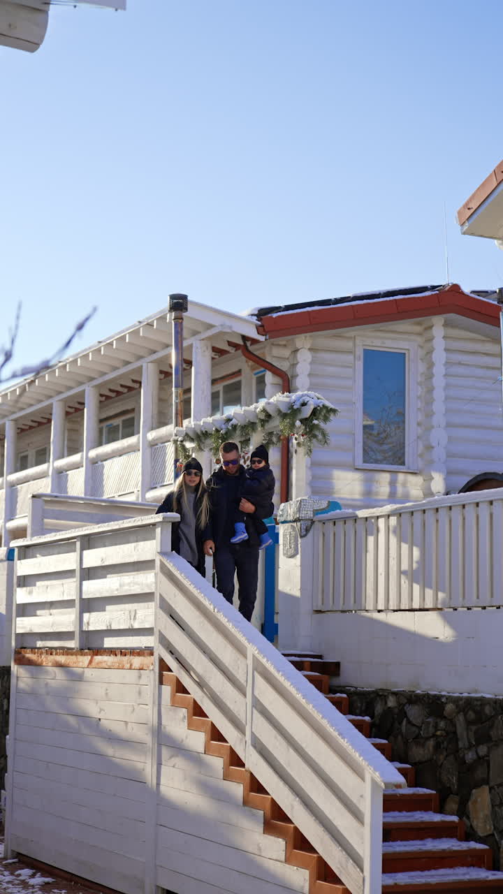 Caucasian parents of a little baby boy stand on the stairs near the cottage. Cheerful people jump joyfully before going down. Vertical video.