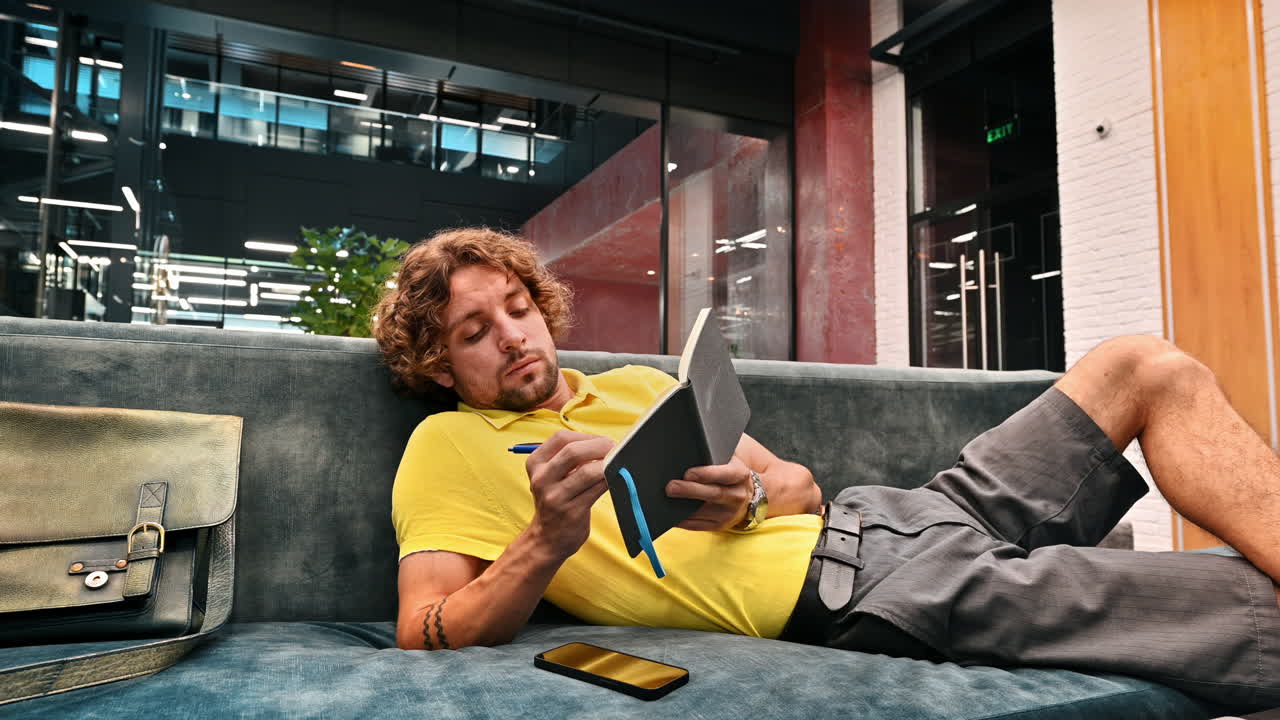 Man in yellow shirt lying on a couch and writing in a notebook