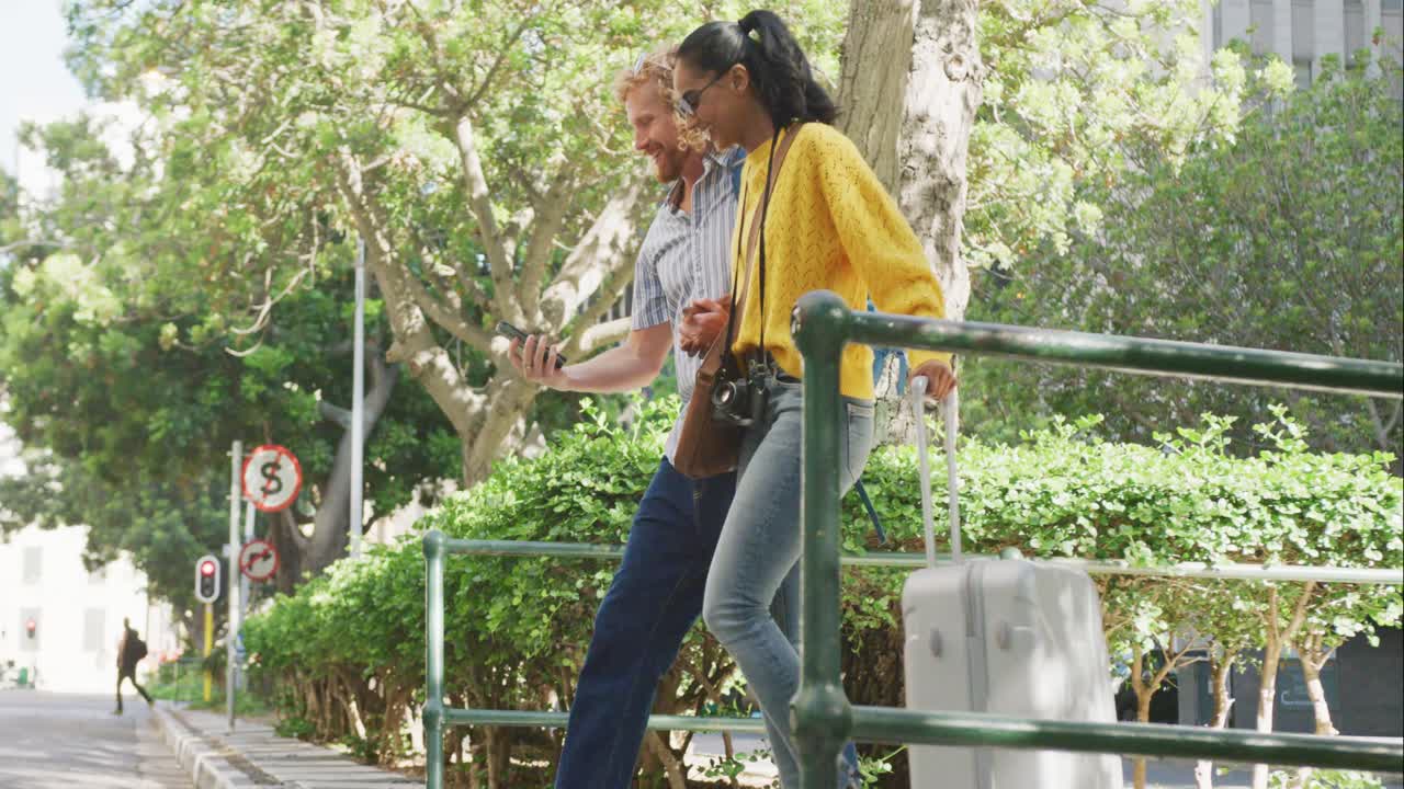 una pareja feliz y diversa caminando con equipaje y usando teléfonos inteligentes en la ciudad.