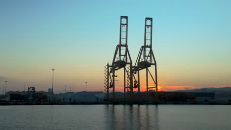 industrial port cranes silhouetted against a serene sunset in málaga, spain. the scene captures the quiet ambiance of an active harbor at dusk