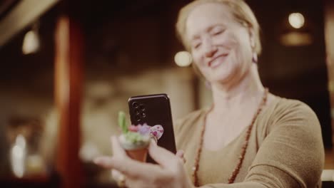 a smiling woman takes a picture of a small plant with her smartphone