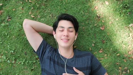 asian man relaxing with eyes closed listening to music on comfortable grass.