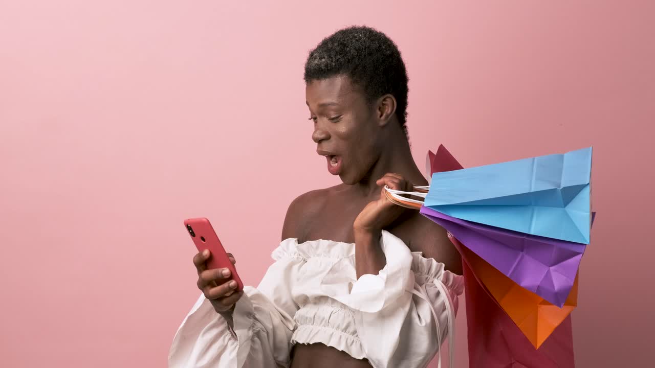 Afrikanische Transgender-Person benutzt ein Mobiltelefon und lächelt, während sie/er Einkaufstaschen hält.